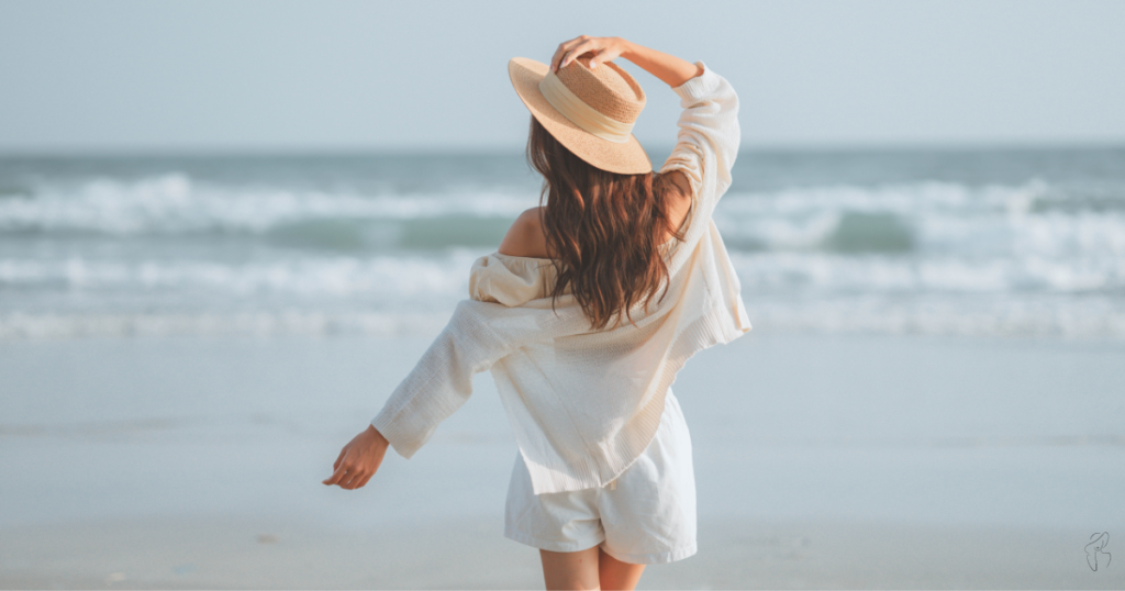 A stylish woman in a white shirt and hat, enjoying the beach with a serene backdrop of sand and sea.